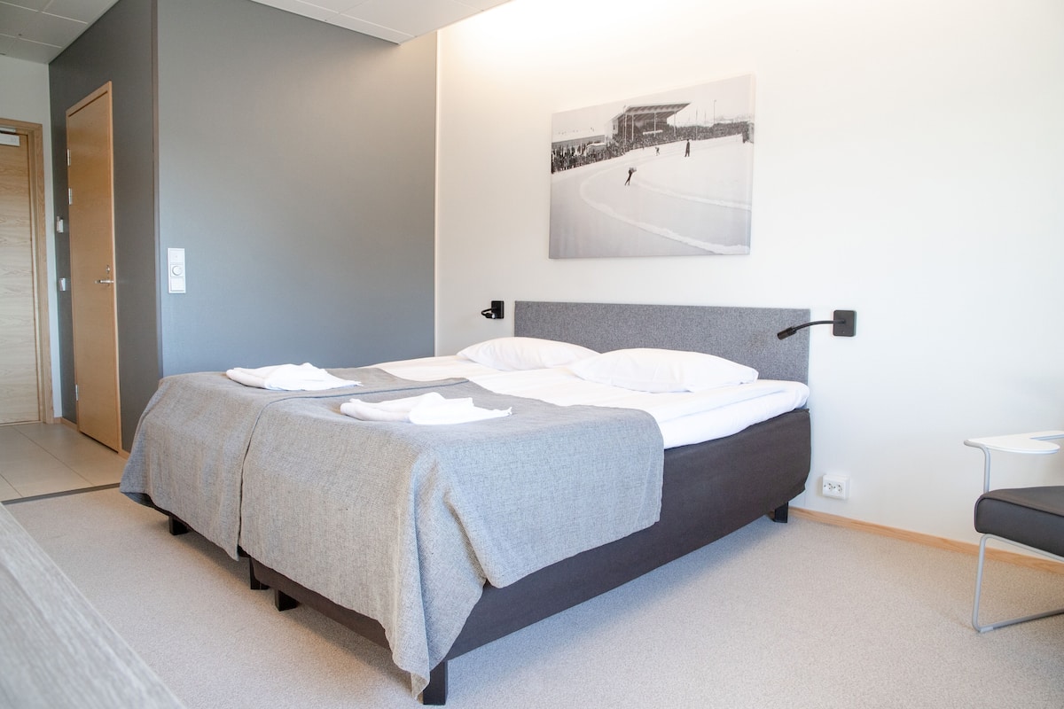 A neatly made bed with a grey blanket and white pillows, a small bedside table, and a wall-mounted picture of a snowy scene.
