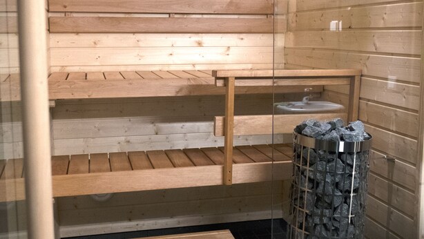 Een sauna met houten banken en een glazen afscheiding, een metalen asbak en een betegelde vloer.