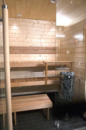 A sauna with wooden benches and a glass partition, a metal ashtray, and a tiled floor. - Santasport Resort (Rovaniemi)