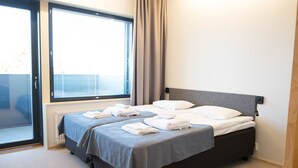 Een netjes opgemaakt bed met witte lakens en een grijze deken in een hotelkamer met een groot raam en een radiator.