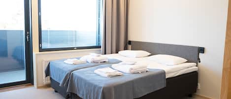 A neatly made bed with white linens and a grey bedspread in a hotel room with a large window and a radiator.