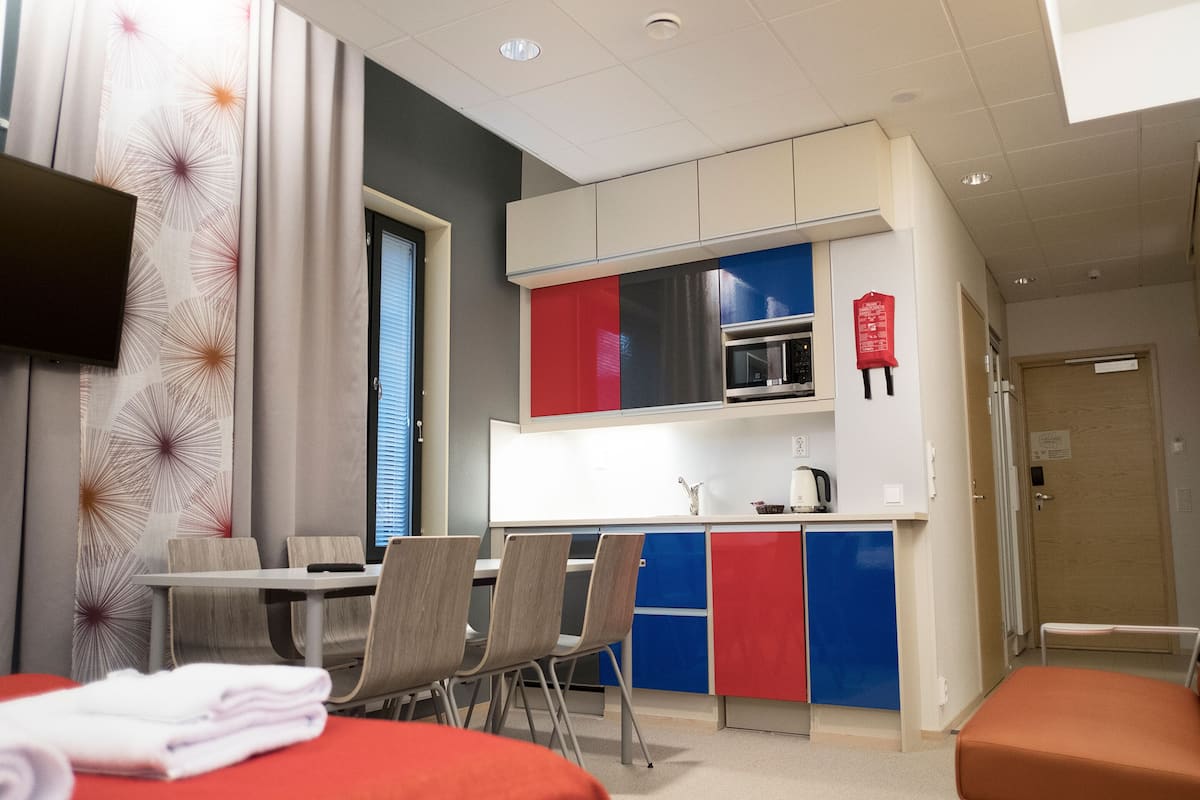 A modern hotel room with a kitchenette, a dining area with chairs, a flat-screen TV, and a bed with a red cover.