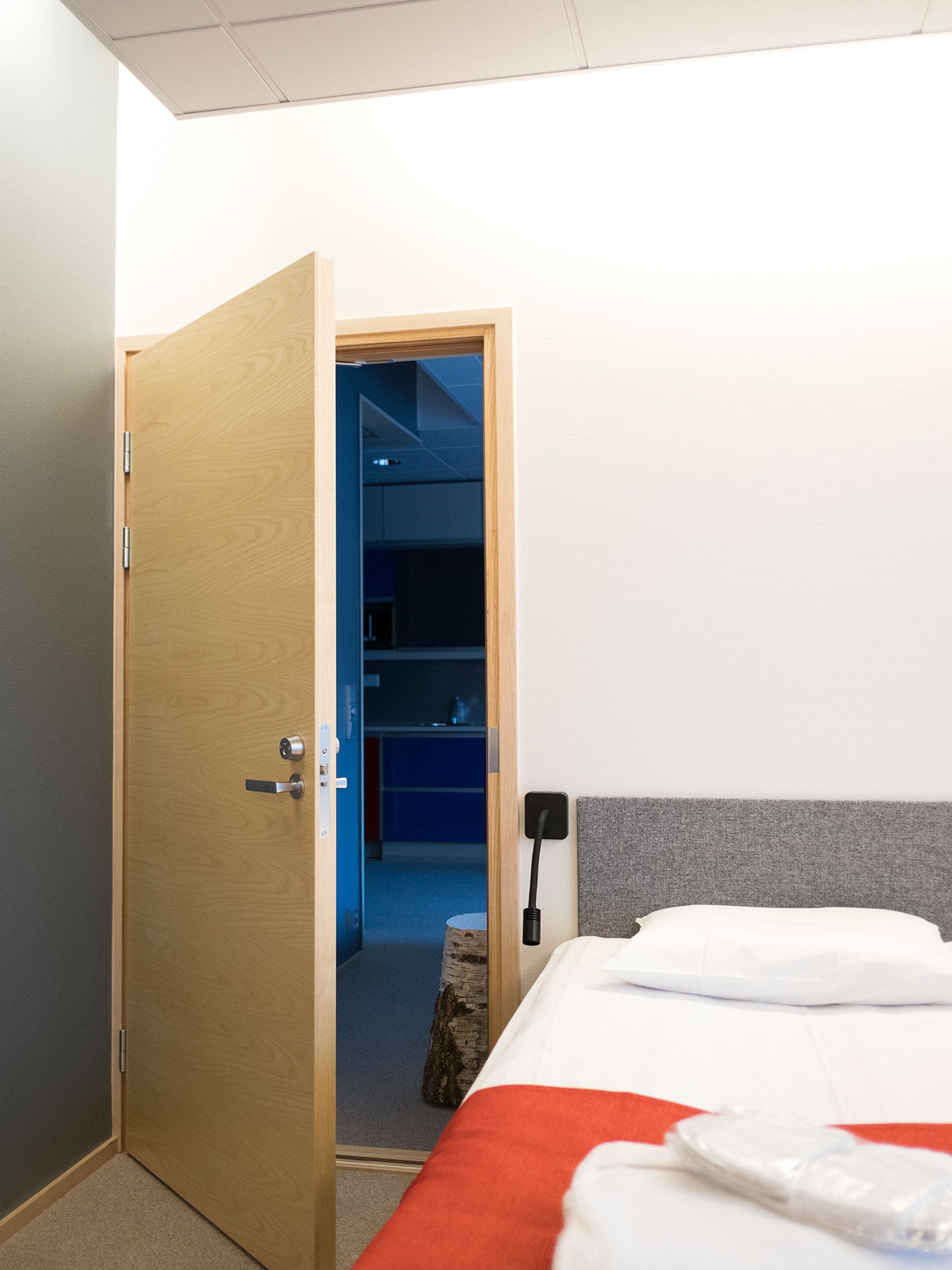 Une chambre d’hôtel avec une porte en bois, un lit avec une literie blanche, un plaid rouge, une table de chevet et une prise électrique au mur.