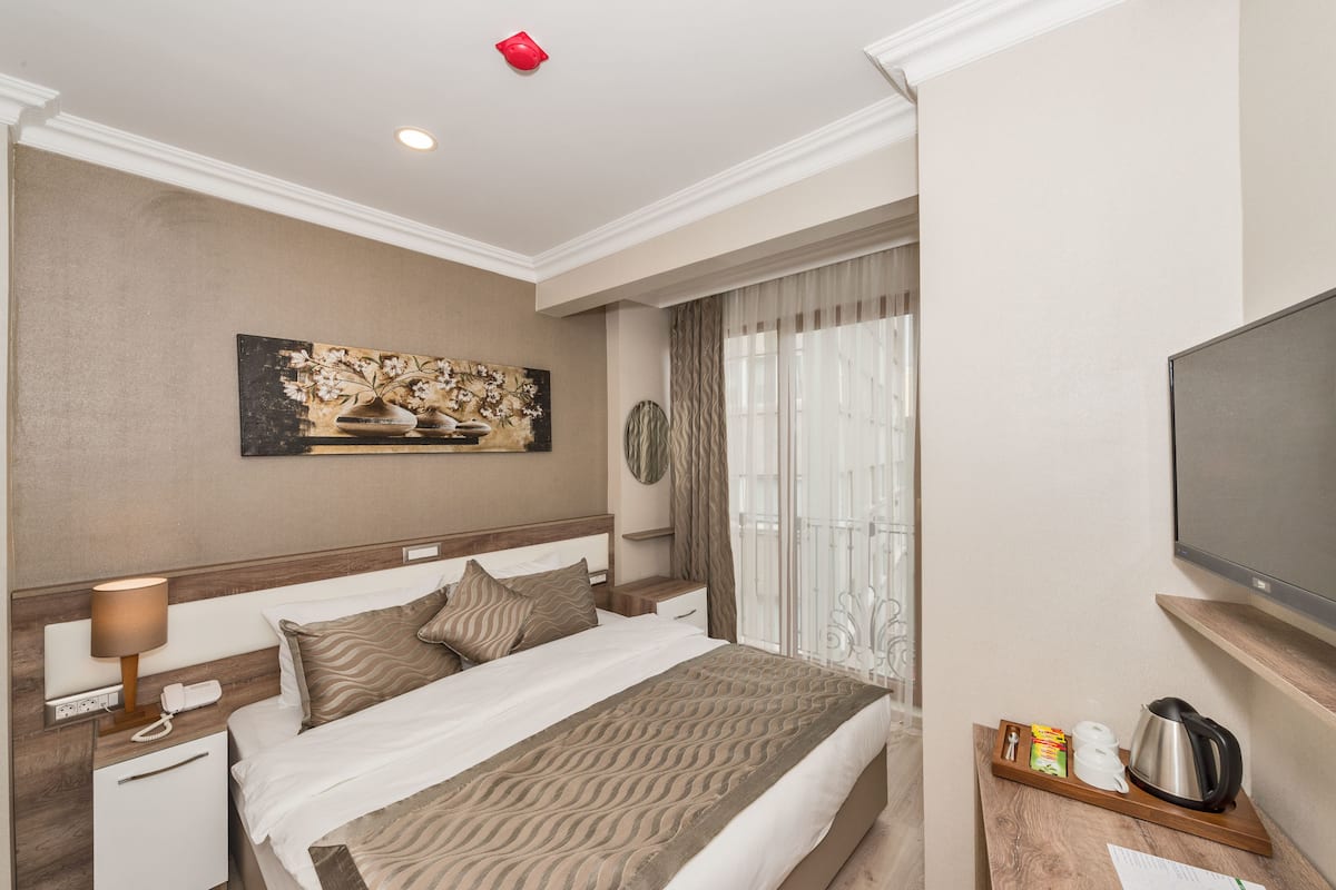 A hotel room with a bed, bedside table, lamp, artwork, curtains, TV, and a tray with a kettle and cups.