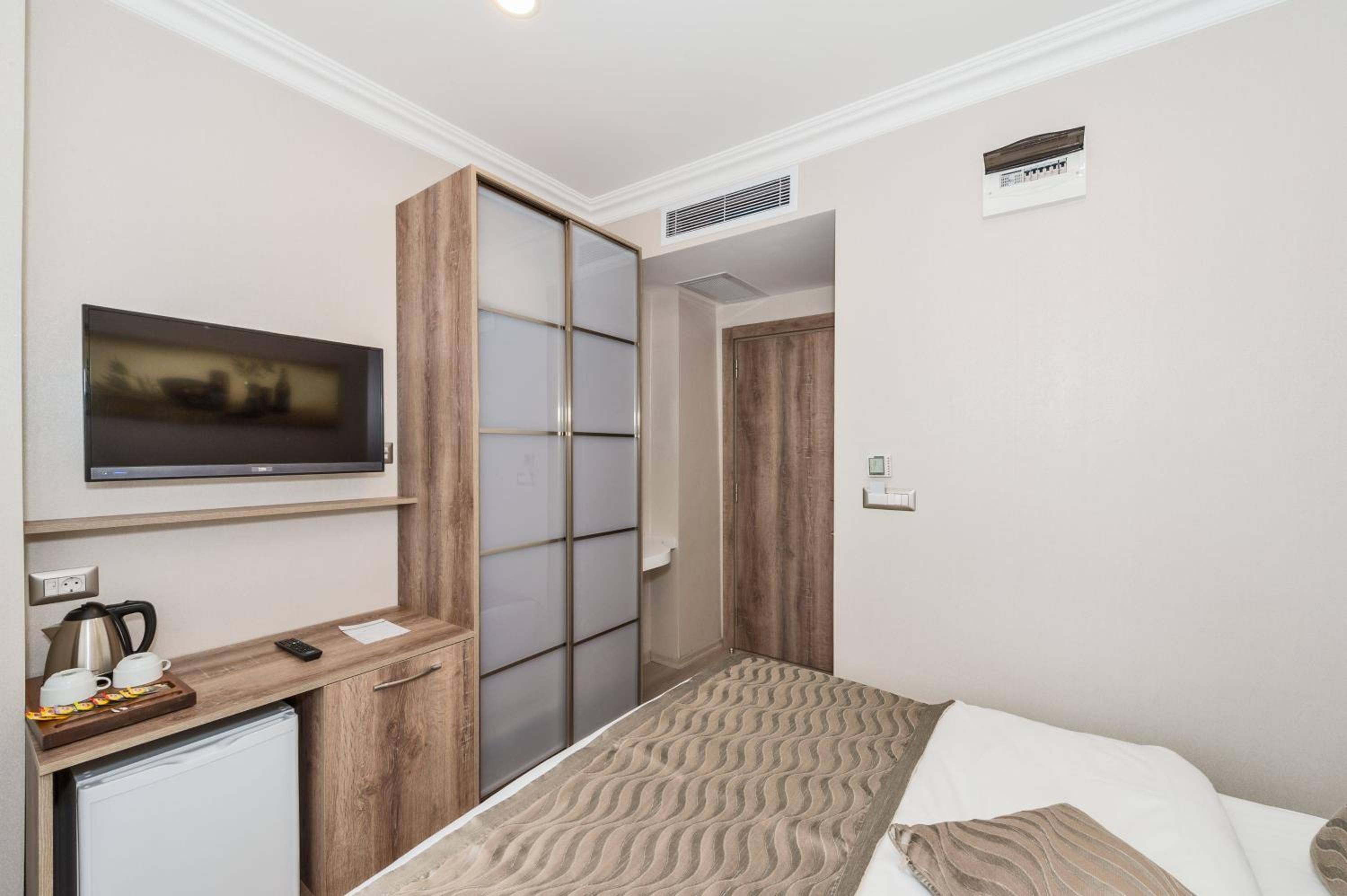 A hotel room with a bed, a TV mounted on the wall, a small kitchenette with a coffee maker, and a sliding door closet.