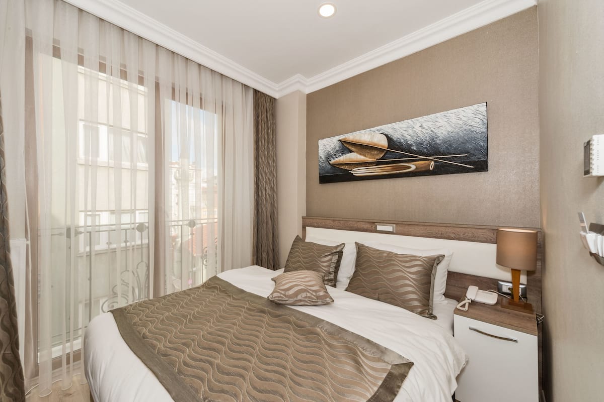A hotel room with a large bed, a nightstand with a lamp, a painting on the wall, and a window with sheer curtains.