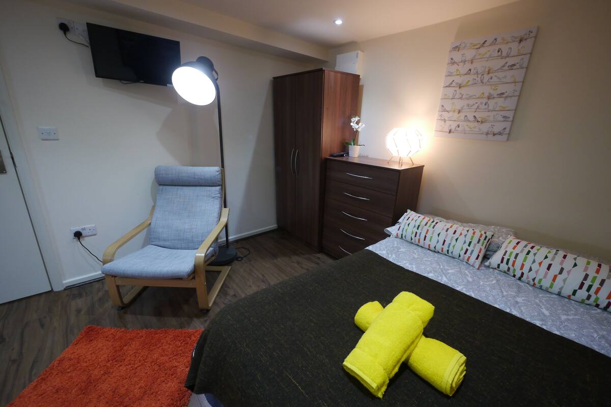 A bedroom with a bed, a chair, a wardrobe, a flat-screen TV, and a wall-mounted light.