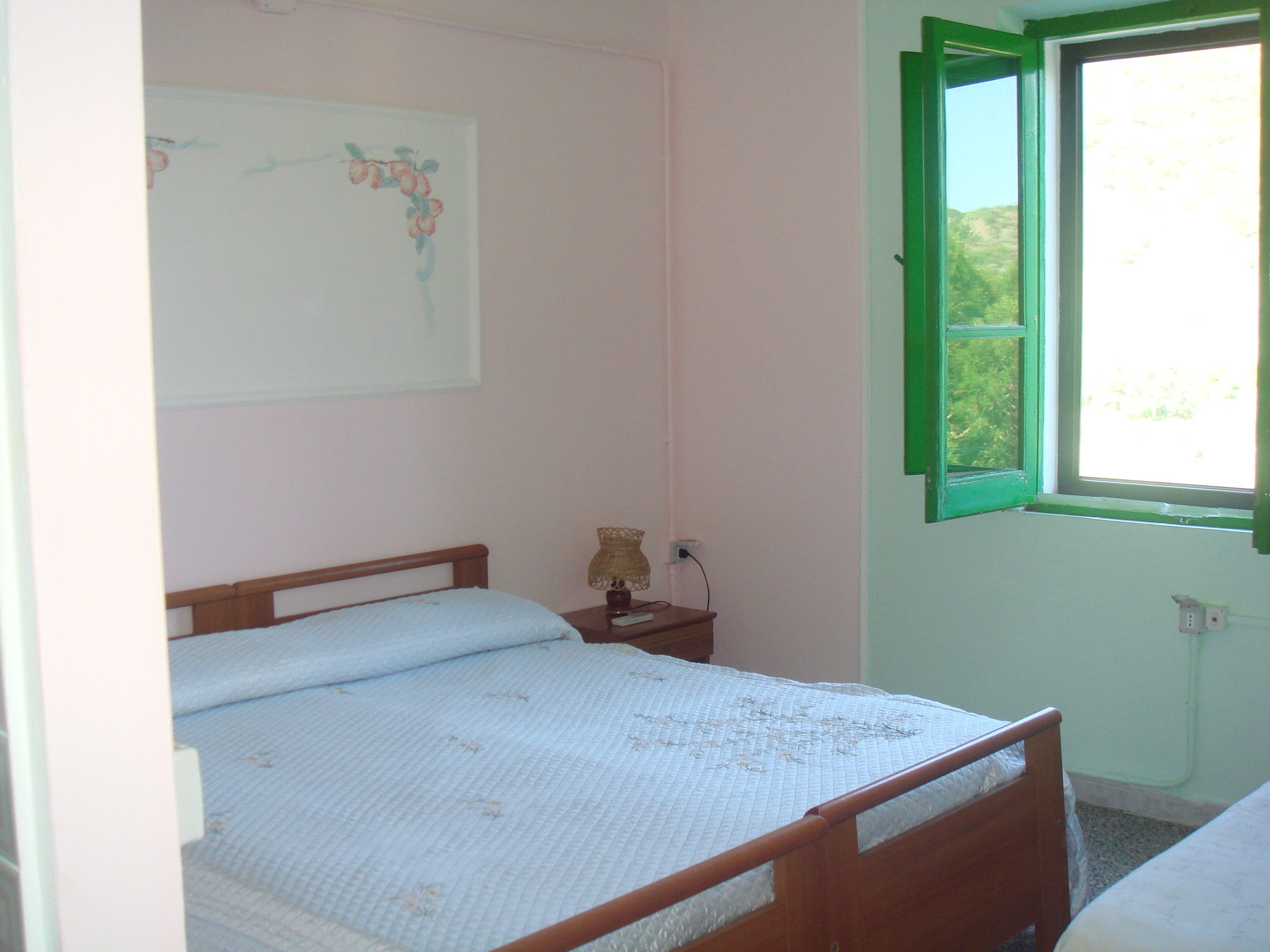 A small, simple bedroom with a single bed, a nightstand, a lamp, and a window with green shutters.