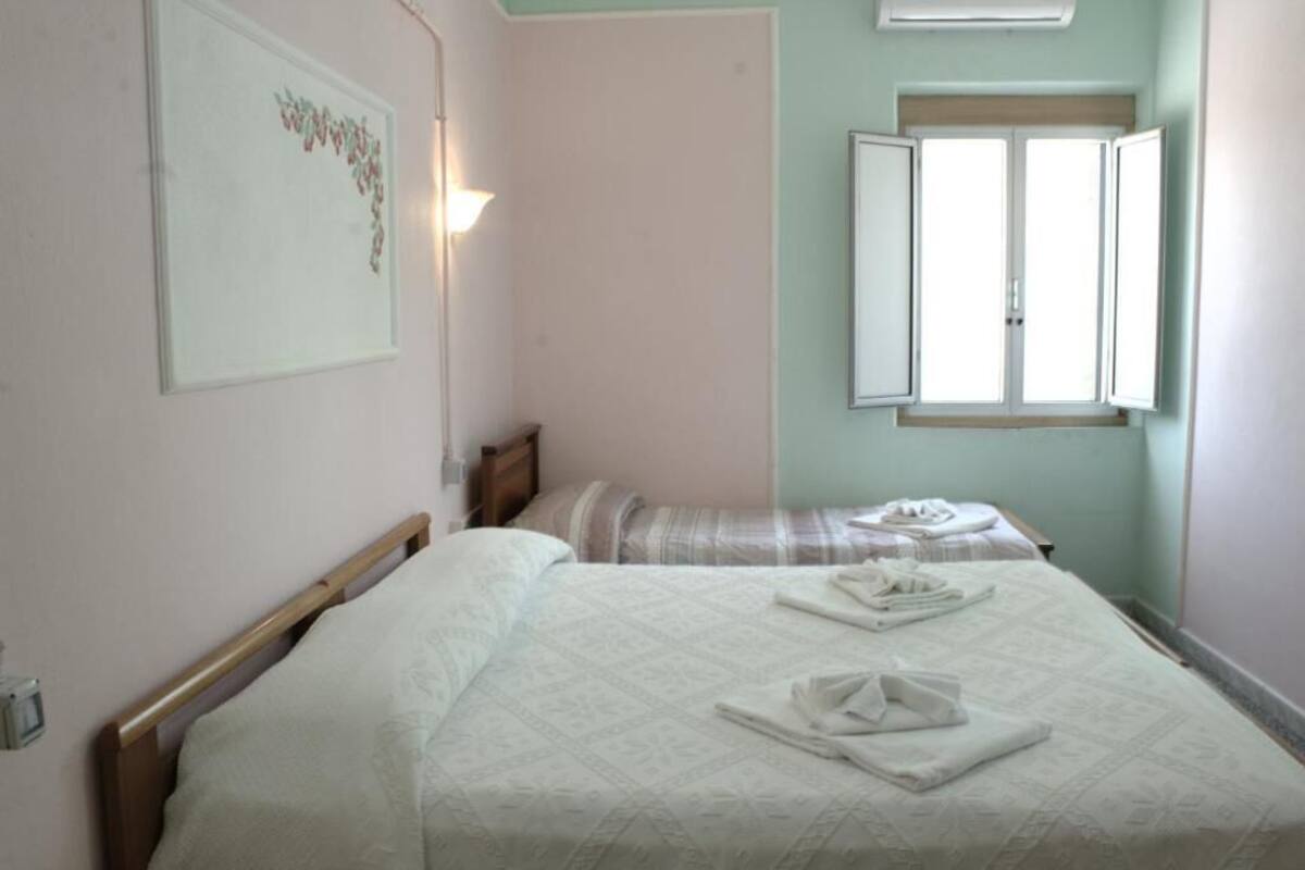 Un dormitorio con dos camas, una mesita de noche, una lámpara de pared y una ventana con contraventanas abiertas.