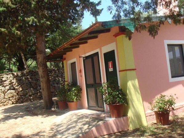 A pink building with a stone wall, two potted plants, and a fire exit sign. - Agriturismo Menhir (Villagrande Strisaili)