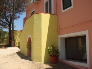 A building with a yellow and pink facade, a visible window, and a potted plant. - Agriturismo Menhir (Villagrande Strisaili)