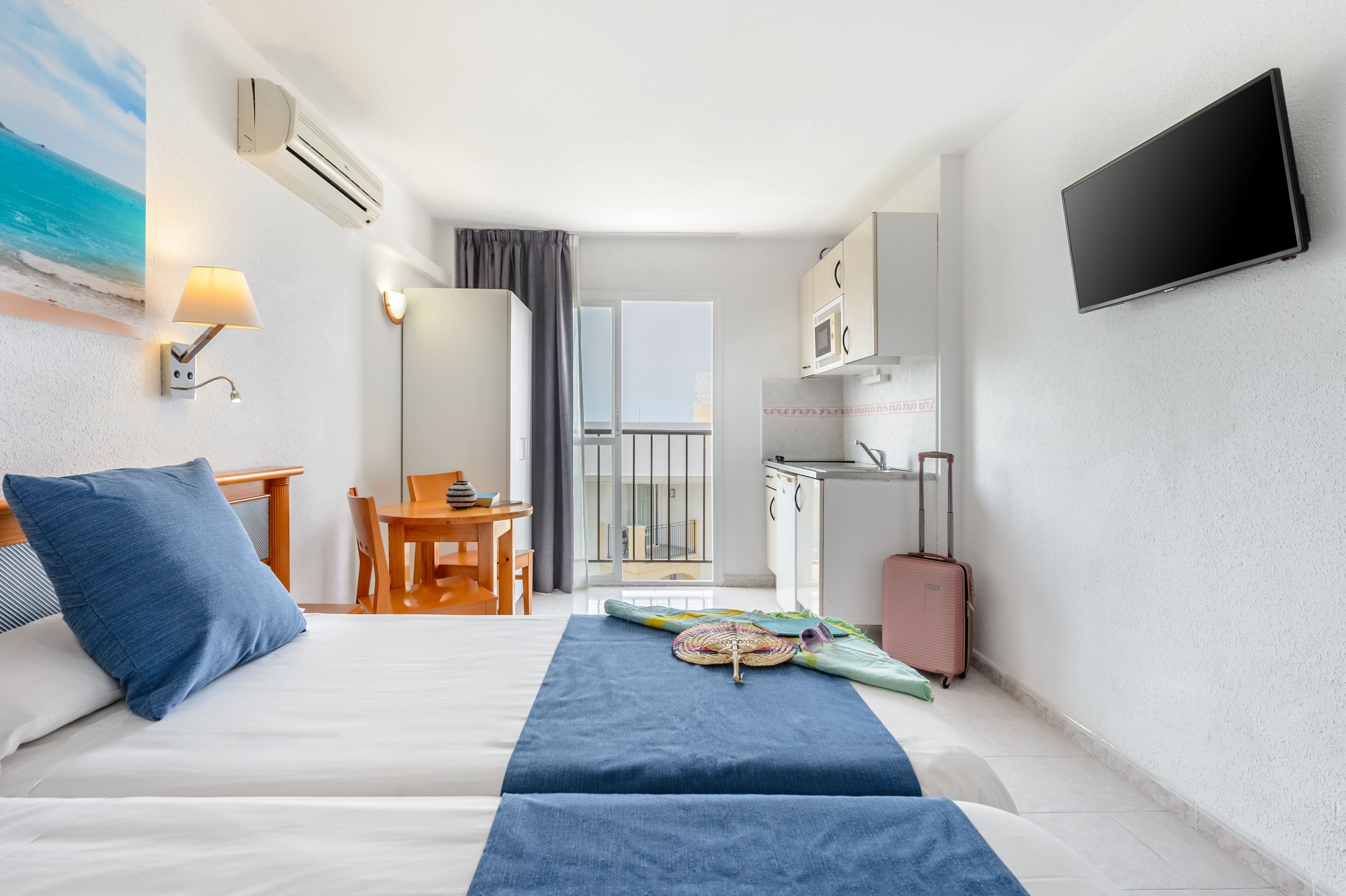 A hotel room with a bed, a desk, a TV, a kitchenette, and a view of the sea.