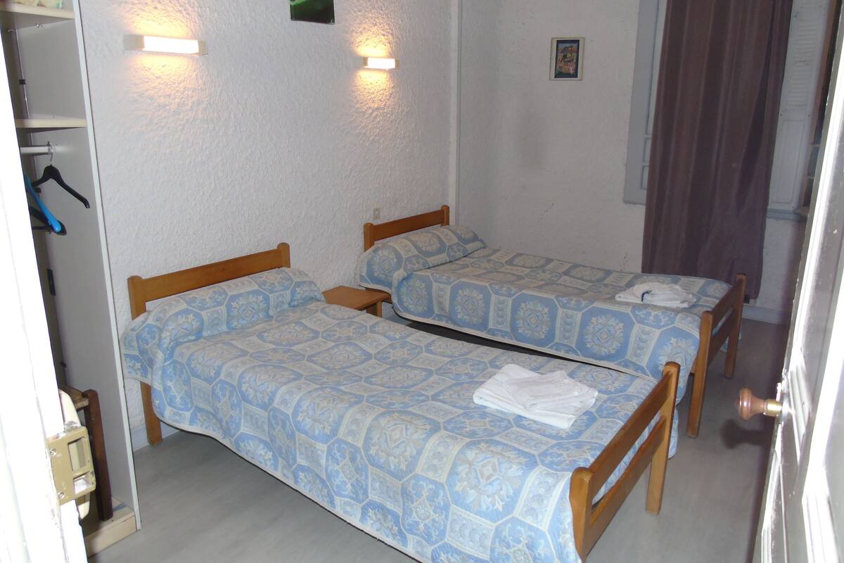 A room with two beds, each with a patterned bedspread, a small bedside table, and a wall-mounted light fixture.