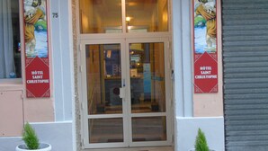 The entrance to Hotel Saint Christophe with a glass door, potted plants, and two red banners with a religious figure. - Hôtel Saint Christophe (Lourdes)