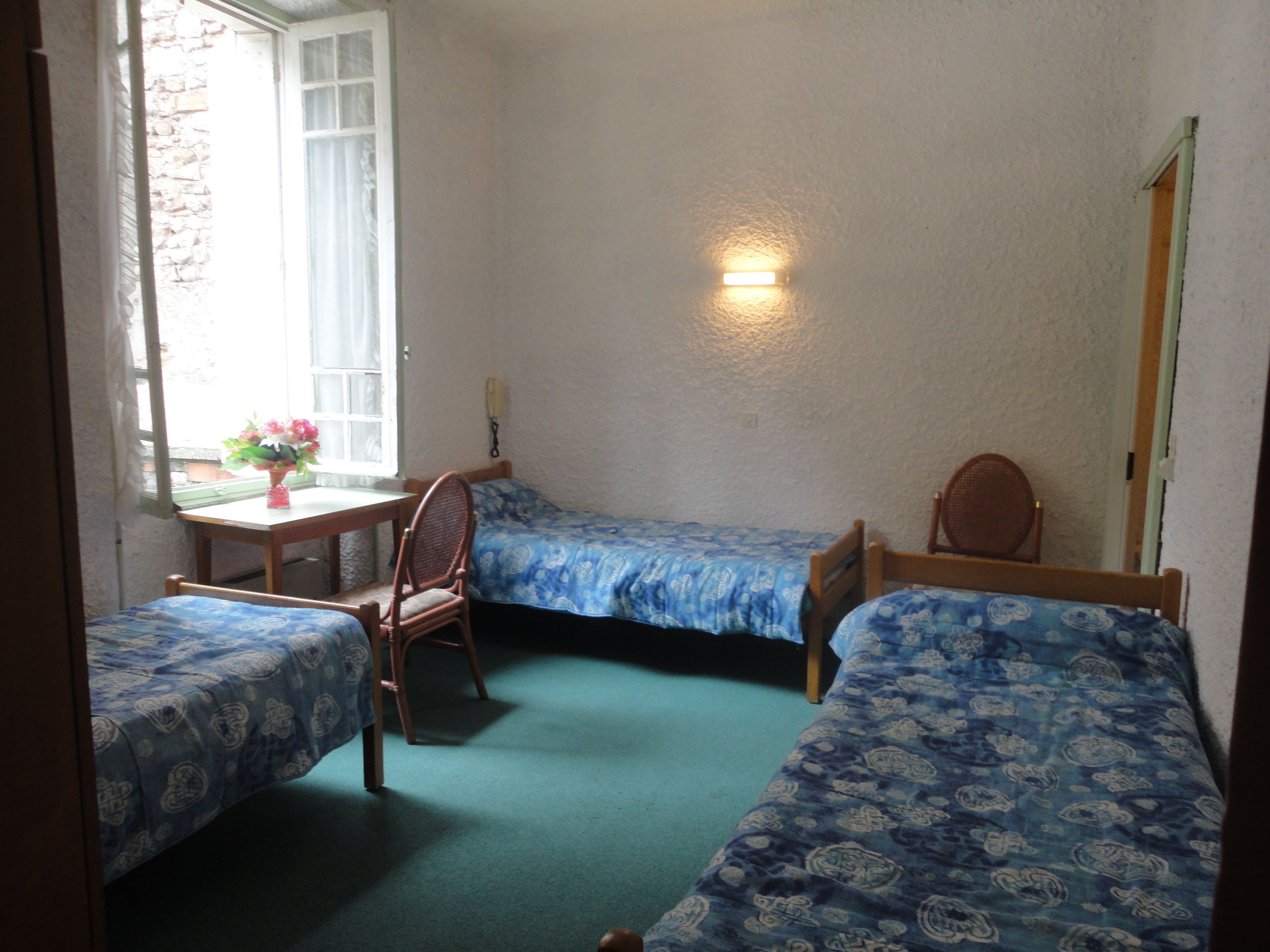 A room with two single beds, a small table with a vase of flowers, a chair, and a window with curtains.
