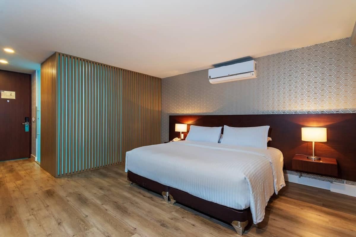 A hotel room with a large bed, wooden flooring, a vertical panel wall, a patterned wallpaper, and an air conditioning unit.