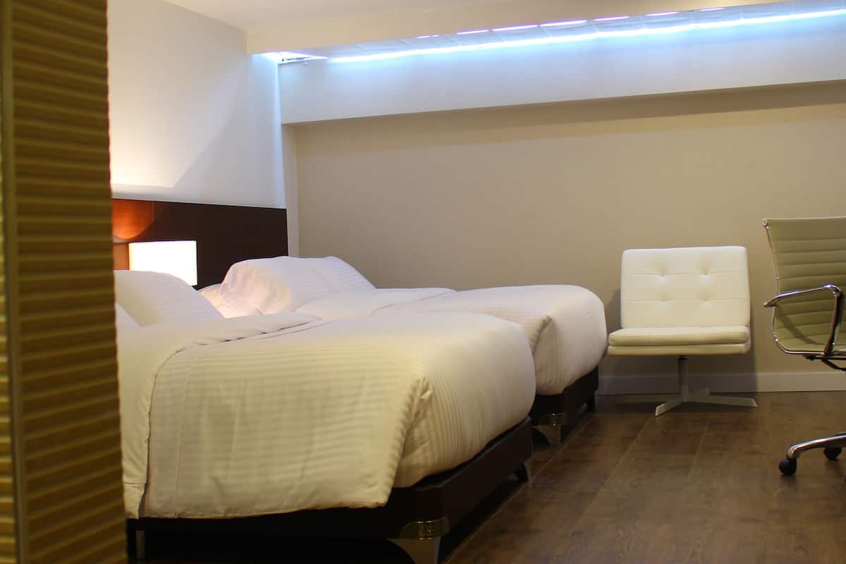 A hotel room with a wooden floor, a bed with white linens, a white armchair, and a desk with a chair.