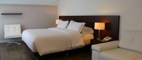 A modern hotel room with a large bed, two bedside tables with lamps, a white armchair, and a sofa.
