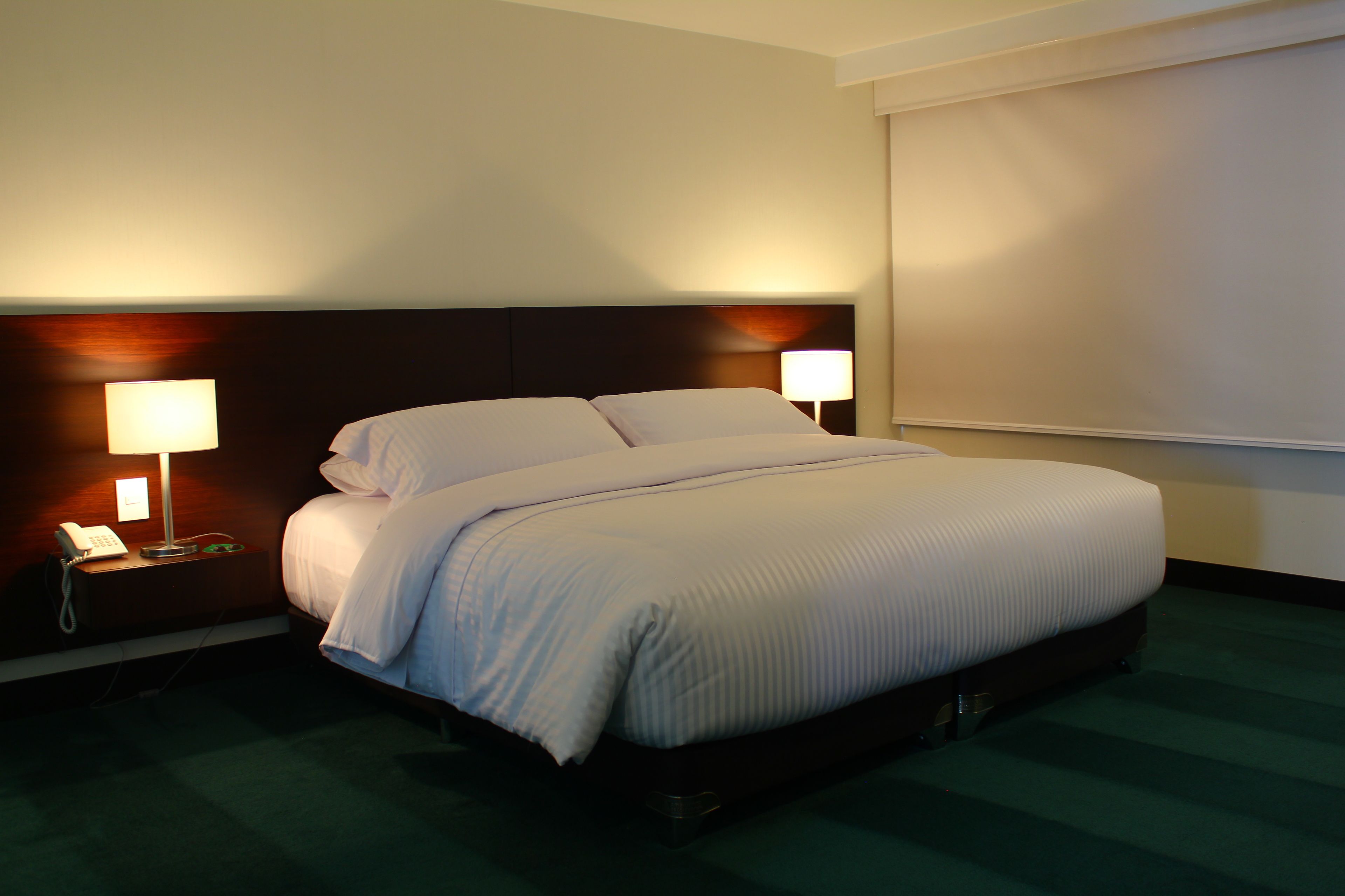 A hotel room with a large bed, two bedside lamps, a telephone, and a window with blinds.