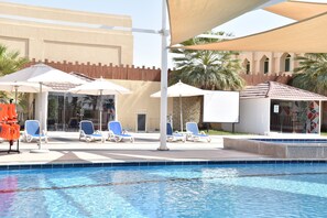 A pool area with lounge chairs and umbrellas, a tiled pool, and a building with a wooden fence and palm trees in the background.