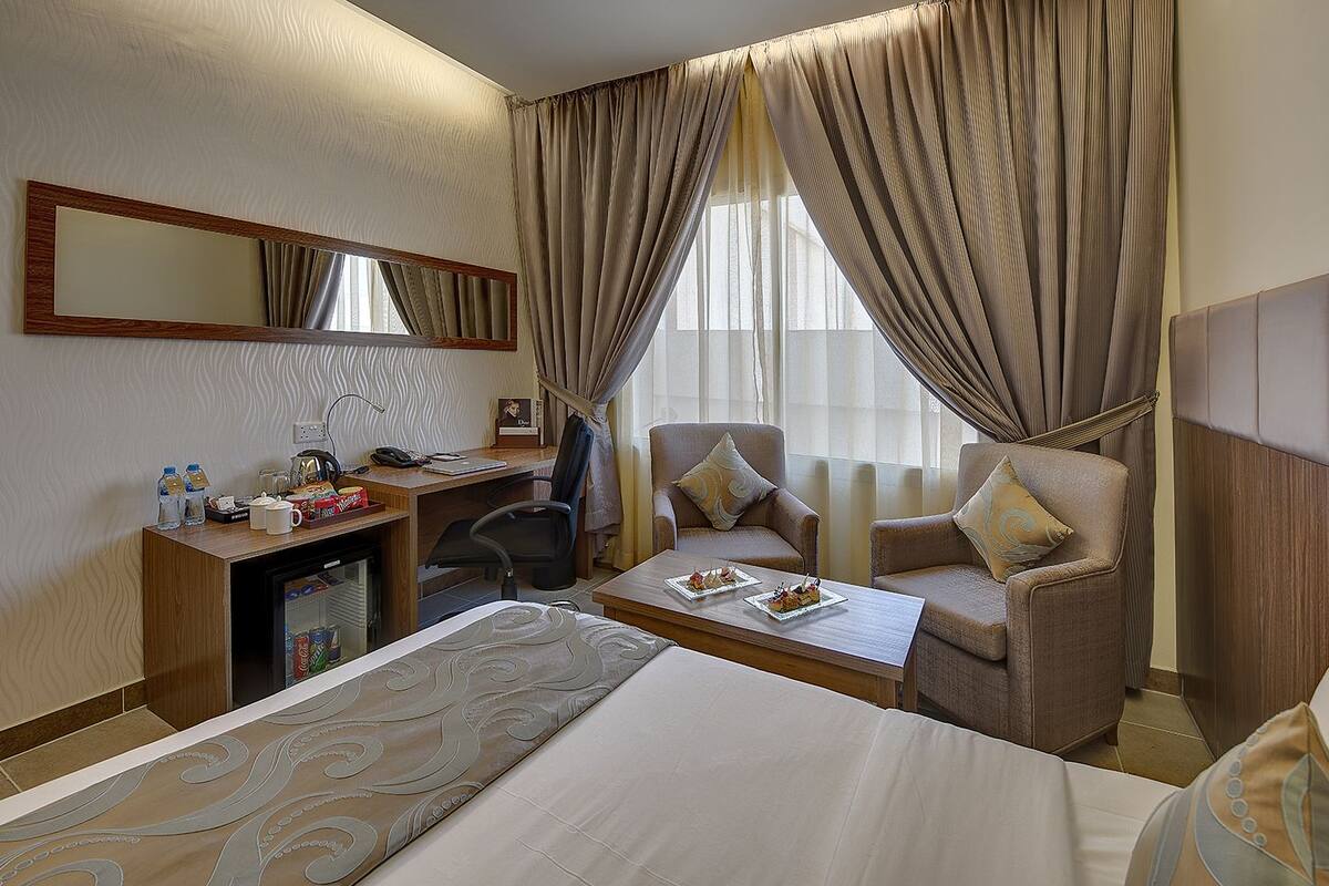A hotel room with a bed, a desk with a computer, a chair, a coffee table with snacks, and a minibar.