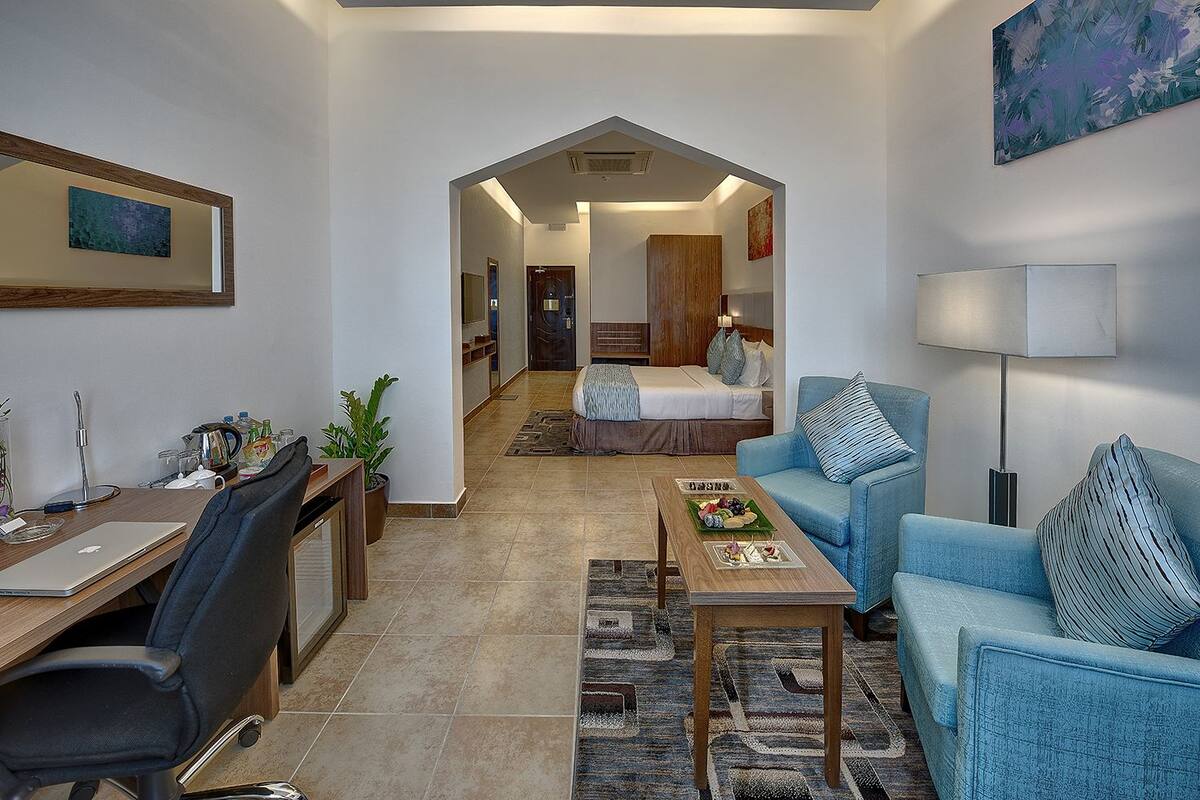 A modern hotel room with a desk, a blue sofa, a coffee table, and a bed in the background.