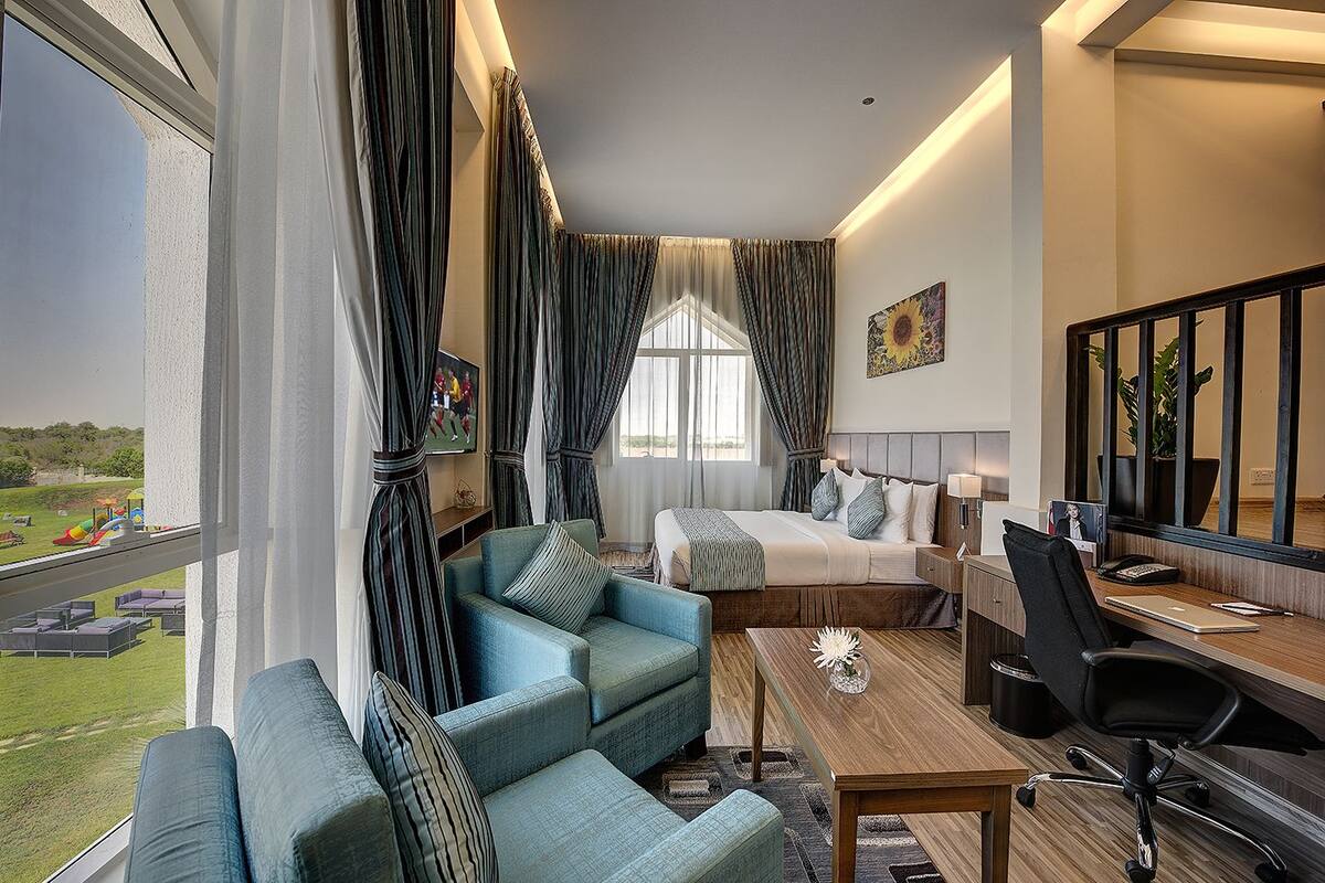A hotel room with a large window, a bed, a desk with a chair, a sofa, and a TV.