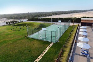 An outdoor basketball court with a glass enclosure, surrounded by a grassy area and a row of sun loungers.