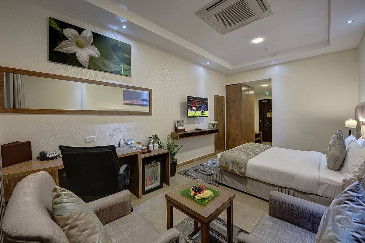 A hotel room with a bed, two armchairs, a desk with a computer, a small refrigerator, a TV, and a wall-mounted mirror.