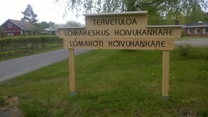 A wooden sign with text in a foreign language, located in a grassy area with a road and buildings in the background. - Koivukankare (Kaarina)