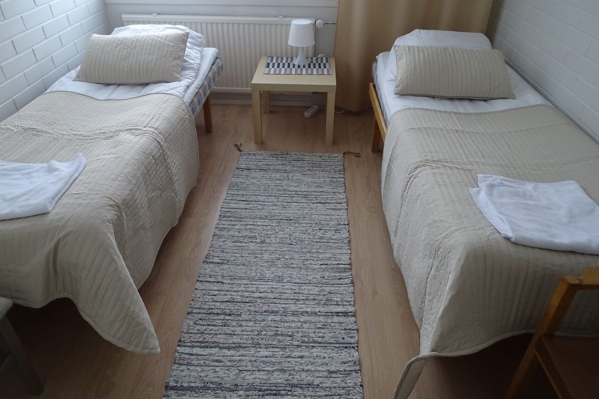 Ein Zimmer mit zwei Einzelbetten, einem kleinen Tisch mit Lampe, einem Fenster mit Vorhängen und einer Heizung.