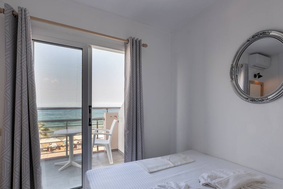 A hotel room with a bed, a large mirror, and a balcony with a view of the sea.