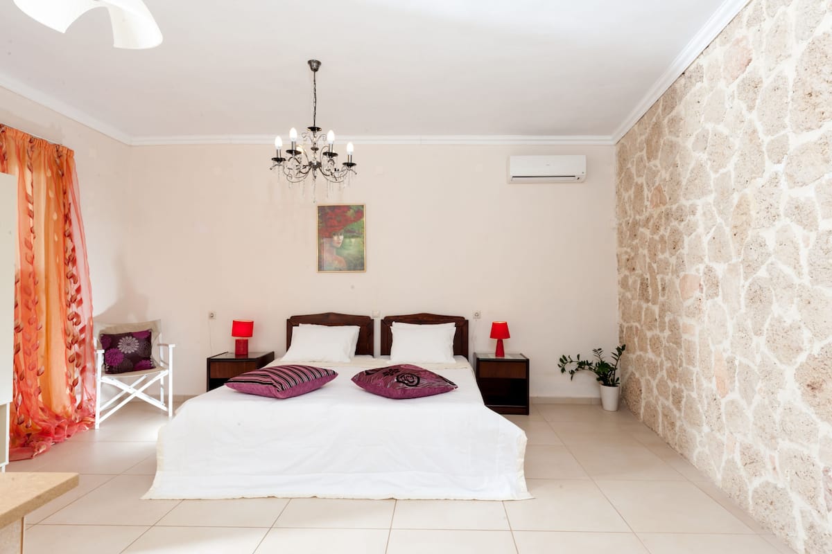 A bedroom with a large bed, two bedside tables with lamps, a chandelier, a painting on the wall, and a stone wall.