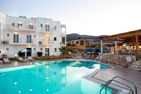 A swimming pool with lounge chairs and umbrellas, surrounded by multi-story buildings with balconies and outdoor seating areas.