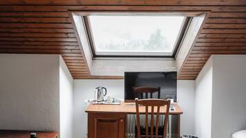Une petite pièce agréable, avec un plafond en pente, une lucarne, un bureau en bois, une chaise et une petite table.