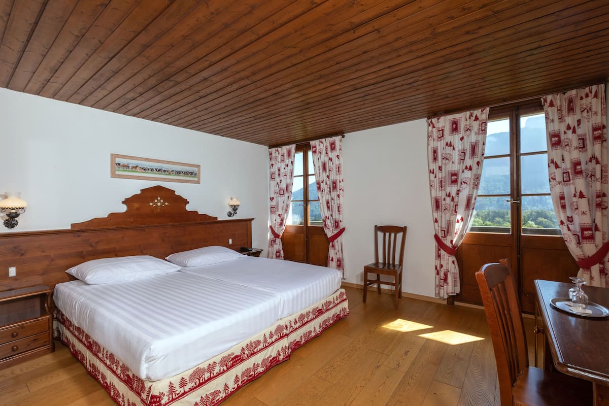 Ein Schlafzimmer mit Holzboden, einem großen Bett mit weißer Bettdecke, zwei Nachttischen mit Lampen, einem Holzstuhl, einem Esstisch mit einem Glas und einem Fenster mit gemusterten Vorhängen.