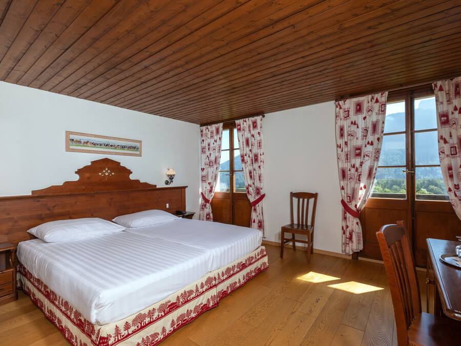 Ein Schlafzimmer mit Holzboden, einem großen Bett mit weißer Bettdecke, zwei Nachttischen mit Lampen, einem Holzstuhl, einem Esstisch mit einem Glas und einem Fenster mit gemusterten Vorhängen.