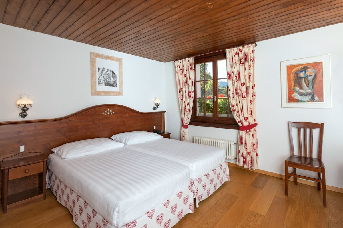 Ein Schlafzimmer mit Holz-Kopfteil, einem Bett mit weißer Bettwäsche, einem Nachttisch, einem Stuhl, einem Fenster mit Vorhängen und zwei Wandlampen.