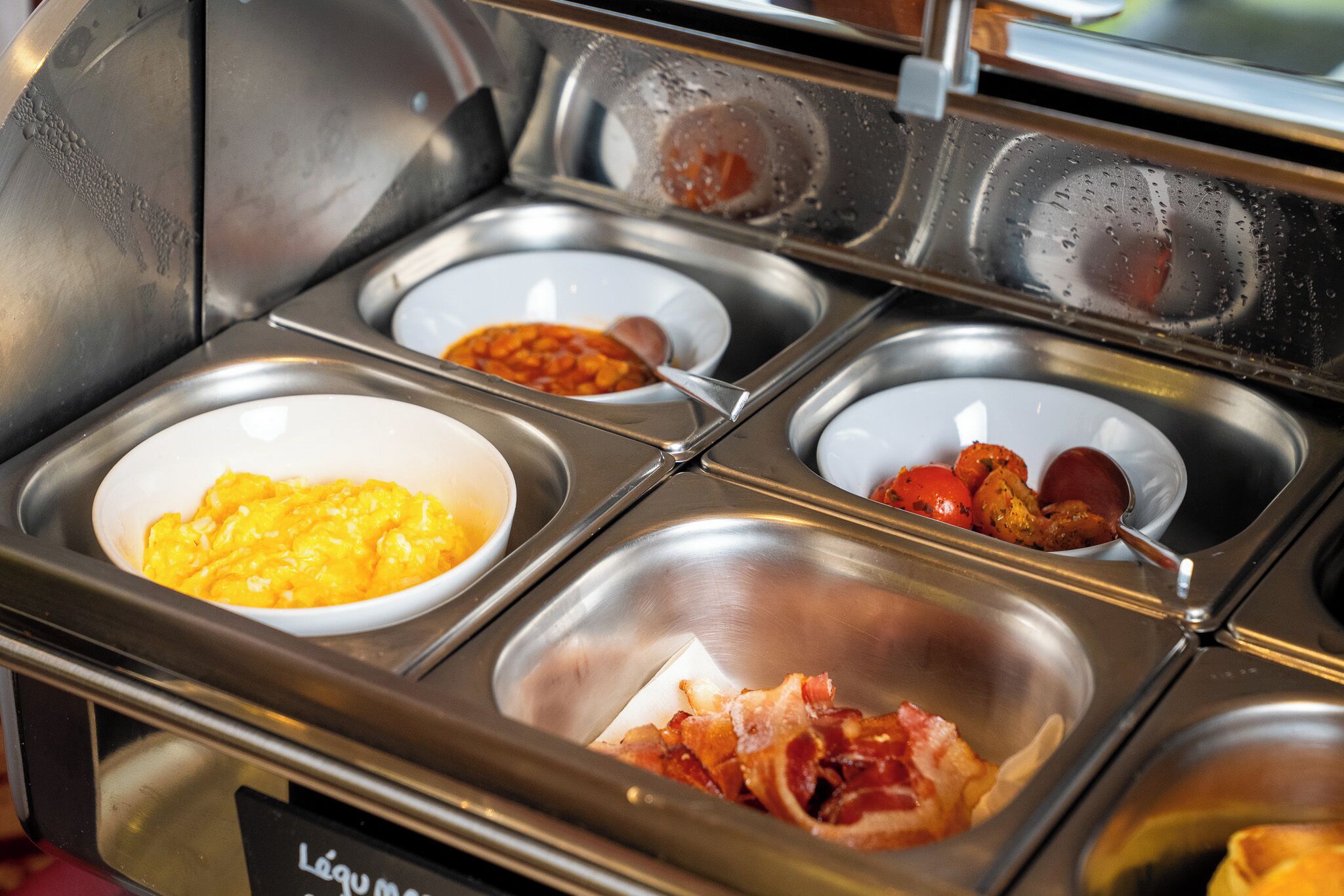 Un bufé con diversas opciones de comida, incluyendo huevos revueltos, tocino y un recipiente con salsa.