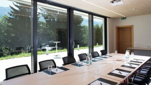 Un salón de conferencias con una larga mesa, sillas y ventanas de vidrio que ofrecen vistas a mesas exteriores y follaje.
