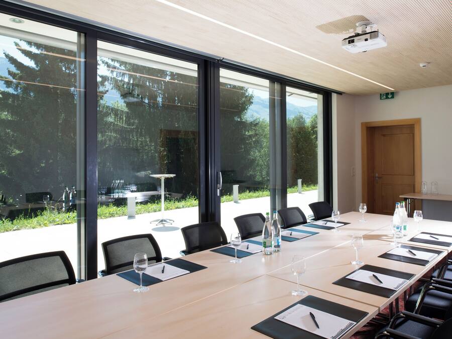 Un salón de conferencias con una larga mesa, sillas y ventanas de vidrio que ofrecen vistas a mesas exteriores y follaje.