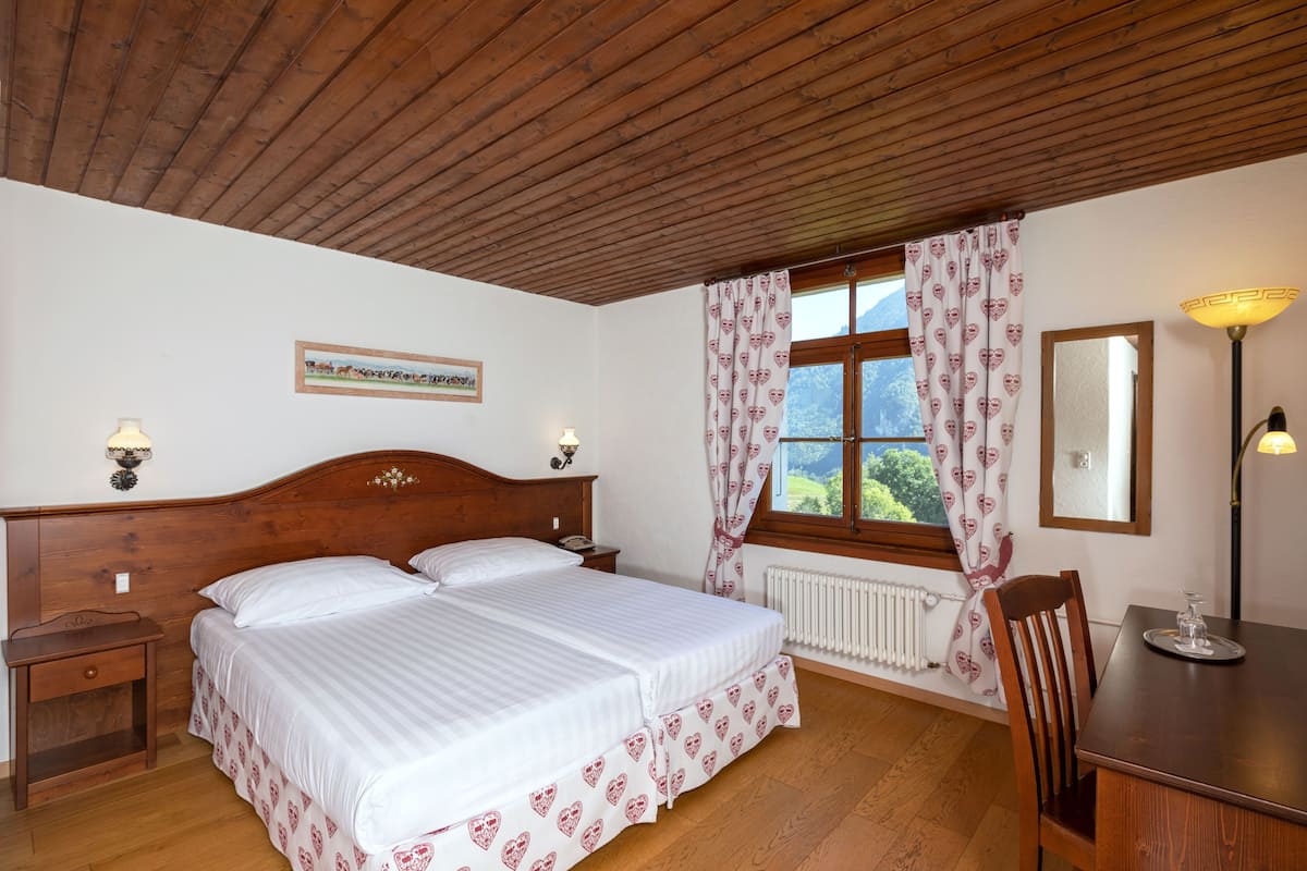 Ein Schlafzimmer mit Holzboden, einem Bett mit weißer Bettwäsche, einem Nachttisch aus Holz, einem Tisch mit Stuhl und einem Fenster mit Vorhängen.
