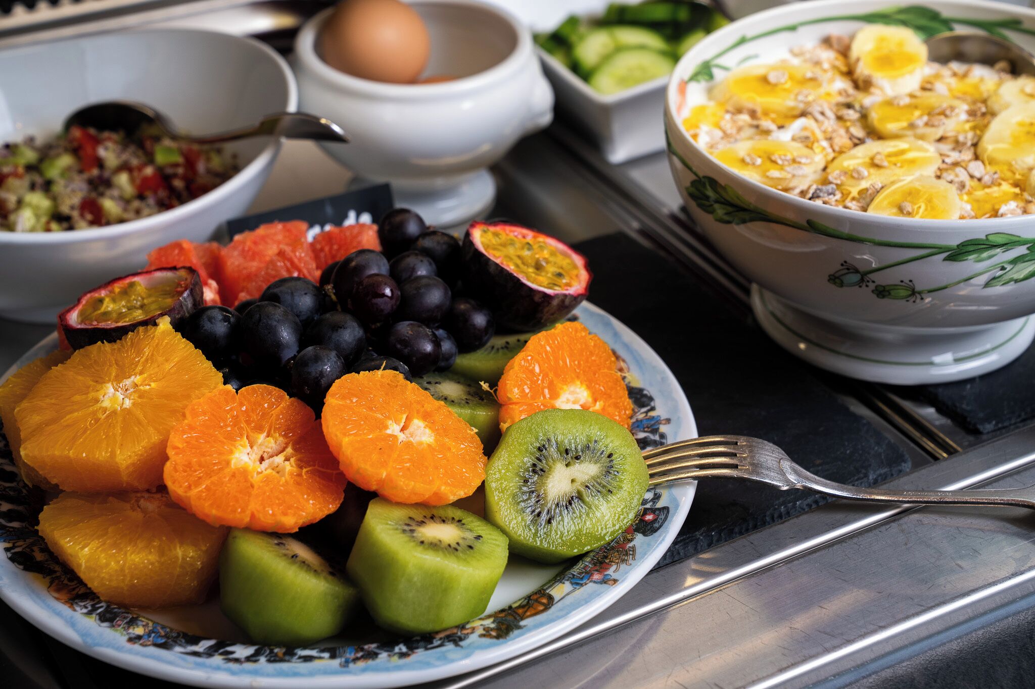Un plato de frutas frescas, como naranjas, kiwi, maracuyá y uvas, con un tazón de cereales y una ensalada al fondo.