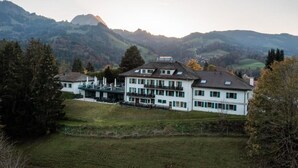 Un edificio grande de dos pisos con techo plano y numerosas ventanas, rodeado de césped y árboles, con montañas al fondo.
