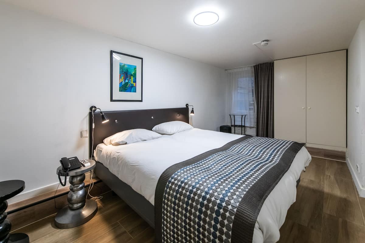 A modern hotel room with a large bed, a bedside table with a phone, a framed artwork on the wall, and a wardrobe.