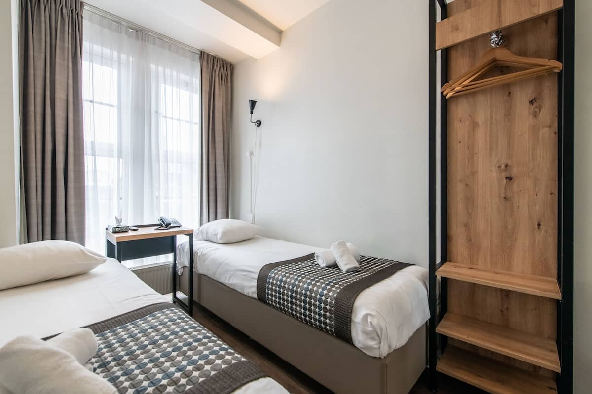 A hotel room with two beds, a wooden wardrobe, a desk with a lamp, and a window with curtains.