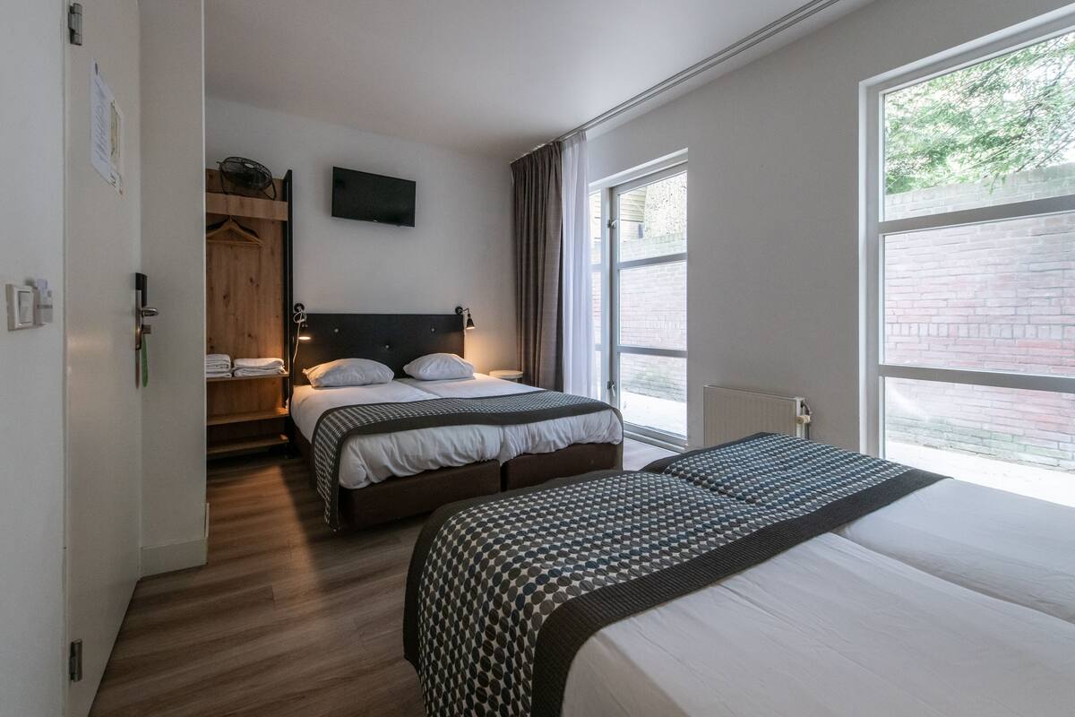A hotel room with two beds, a wooden wardrobe, a flat-screen TV, and large windows.