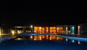 A nighttime view of a hotel with a swimming pool, a covered outdoor area, and a well-lit interior.
