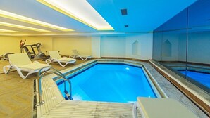 Una piscina interior con agua cristalina y azul, rodeada de sillones de descanso y equipo de ejercicio.