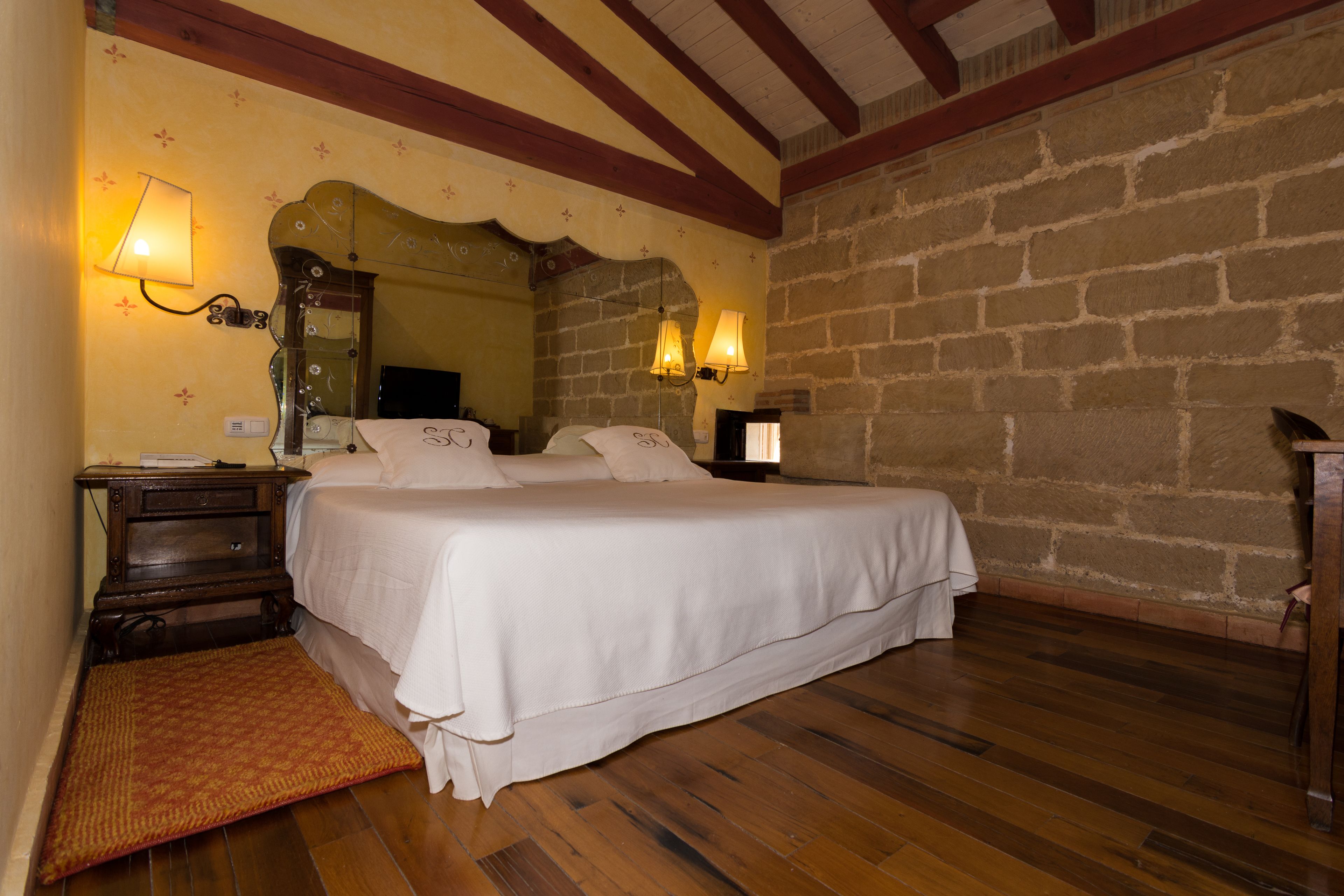 Un dormitorio con una cama grande, suelo de madera, pared de piedra y dos lámparas de pared.